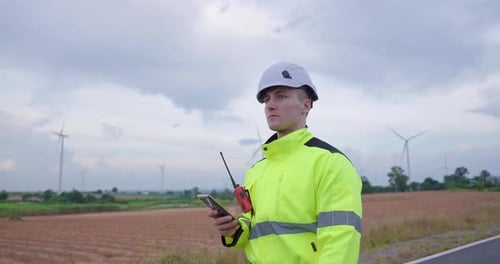 Caucasian engineer holding radio and smartphone near wind farm field, communicating during