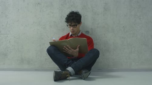 Hispanic male student is sitting in a college hallway and reading a textbook.
