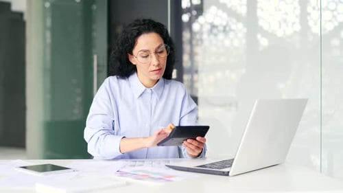 Busy young female accountant forms accounts using laptop and calculator, takes notes while sitting