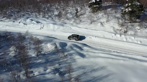 Black van is driving on a snow covered road near the trees on a sunny day, aerial shot in 4k