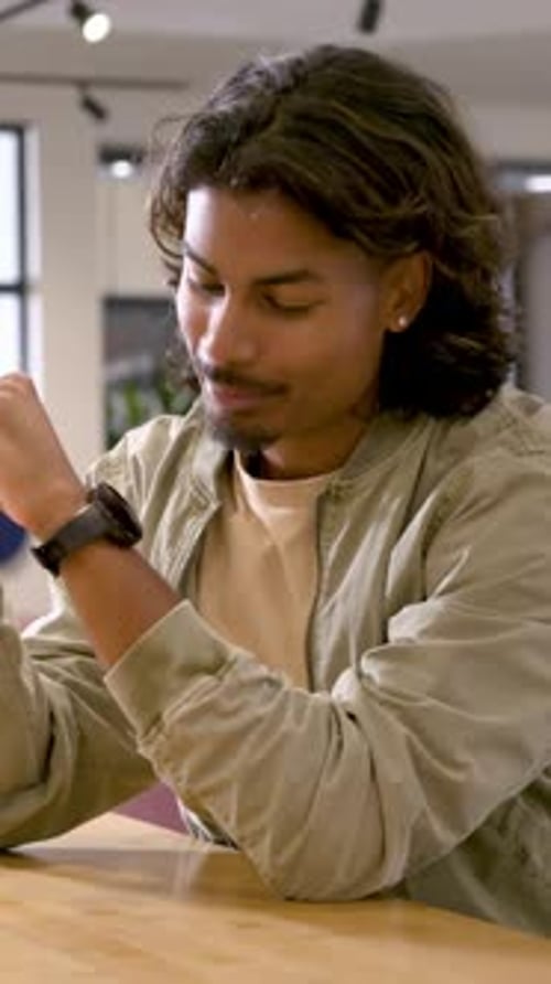 Vertical video: Smiling man in khaki jacket after offscreen prompt, adjusting watch at wooden table