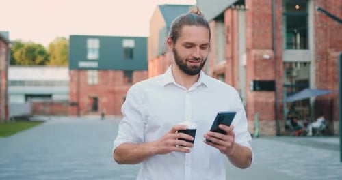 Cheerful bearded businessman walking at street is texting messages and browsing social network.