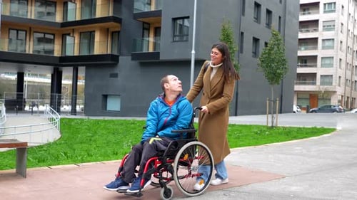 Woman Pushing Man in Wheelchair Along Sidewalk