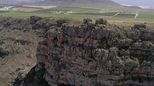 Aerial descends rugged rock escarpment, vineyards, orchards beyond