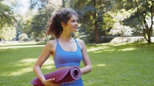 Athletic Fitness Young Woman Walking with Rolled Yoga Mat to Do Fitness Stretching Exercises in Park