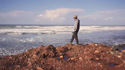Mann mit Hut spaziert am felsigen Strand und schaut auf den endlosen Ozean, friedlicher Reisehintergrund