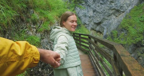 Happy woman holding man hand looking at him and hiking along narrow path in mountains