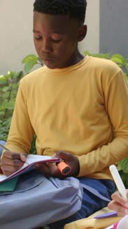 Vertical video: Writing teen boy in yellow shirt in courtyard, holding notebook and orange cylinder
