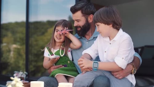 Daughter and Son Sitting on Laps of Dad Little Girl Holding Toy Dragon in Hands Happy Father Talking
