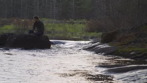 Mann sitzt auf einem Felsen an einem Waldfluss, versunken in Kontemplation, umgeben von Natur und fließendem Wasser