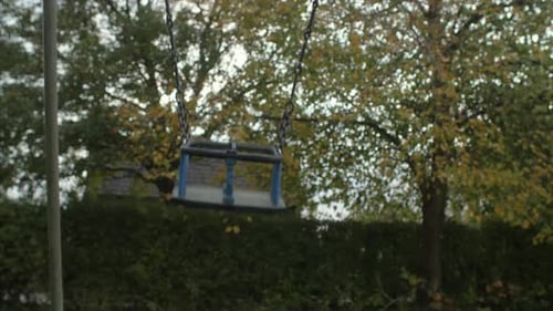 An empty swing in a desolate playground swaying fast. LOW ANGLE FRONTAL VIEW STATIC SHOT.