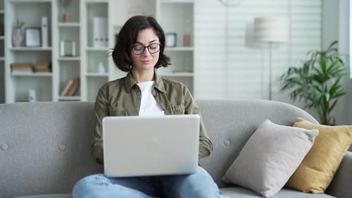 Woman using laptop for remote work at home, seated on comfortable gray sofa. Young female works
