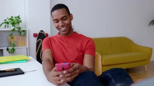Young Happy Millennial Boy Using Mobile Phone Sitting on Desk at Home Studio