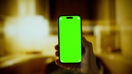 Person Holding Smartphone with Vivid Green Screen in Dimly Lit Indoor Environment Showcasing Blank