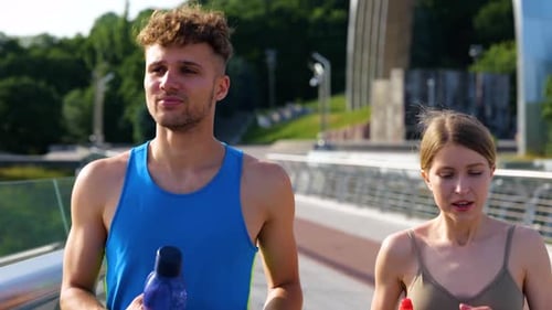 Smiling Man and Woman Running Across the Bridge While Holding Bottles of Water Couple Resting After