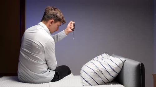 Young Man Sitting in Bedroom Holding Prayer Beads