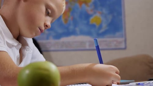 A Schoolboy is Sitting in a Chair at a School Desk and Writing in a Notebook