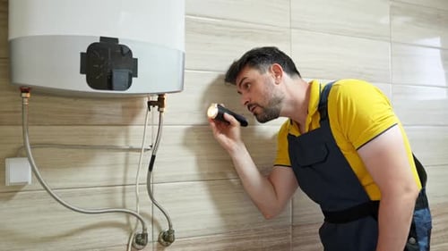 Plumber carefully examining water heater using flashlight, performing maintenance check