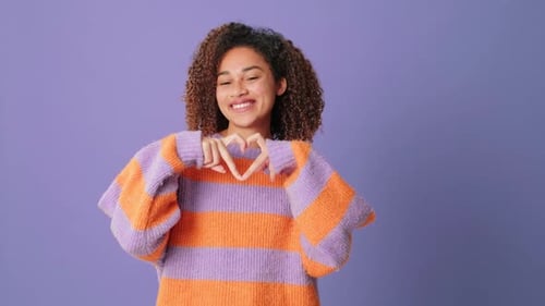 Mujer que muestra un corazón en forma de gesto de dedos mirando a la cámara sobre fondo lila en el estudio