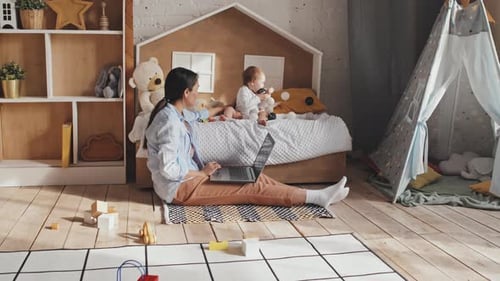 Mom Typing On Laptop on Floor in Nursery Beside Her Baby In Bed