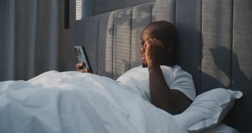 Smiling Man in White Pajamas Relaxes in Bed Holding Smartphone in Hands