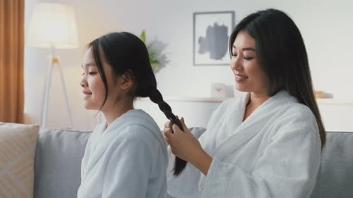 Mother Braids Daughter's Hair While Sitting on the Couch in a Cozy Living Room During the Day