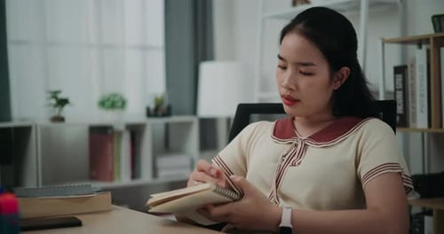 Happy young asian woman sitting at desk while thinking idea holding pen making notes in diary at hom