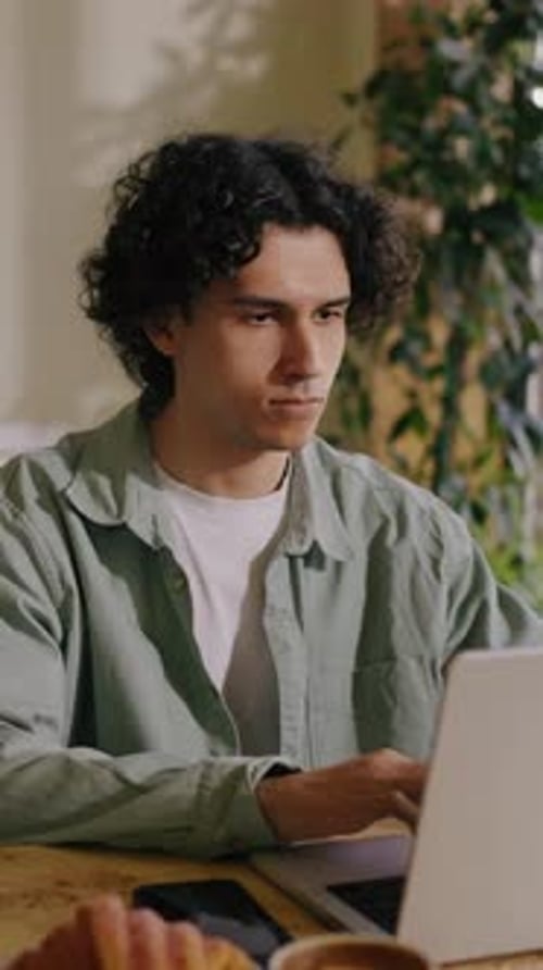 Focused Young Man with Curly Hair Working on a Laptop at a Wooden Table in a Cozy Indoor Space with
