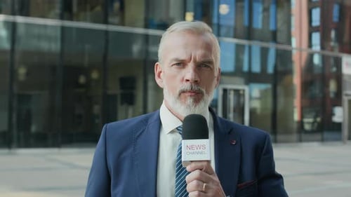Reportero de traje cubriendo noticias frente a un edificio de oficinas de cristal