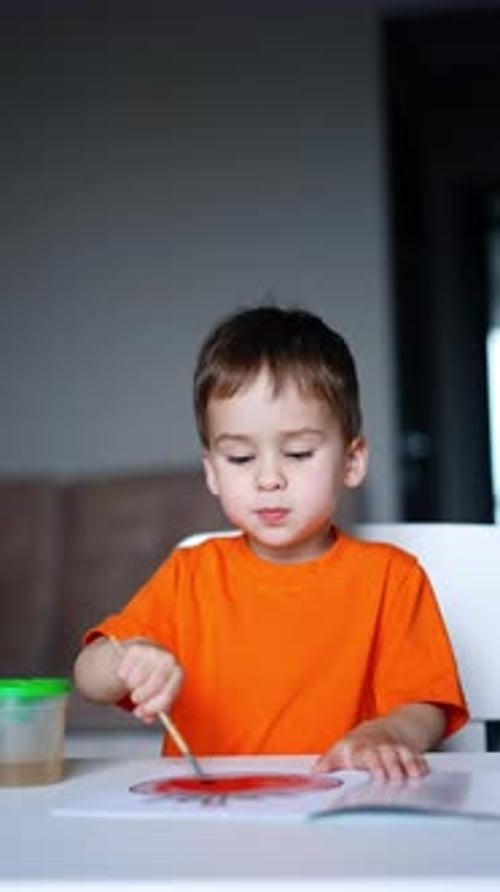 Little baby boy in orange t-shirt sits at desk. Cute toddler is focused on the painting.