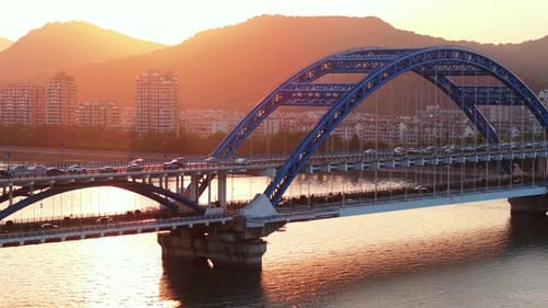 Aerial View of a Blue Bridge with Cars Crossing Over a River at Sunset with a City Skyline in the