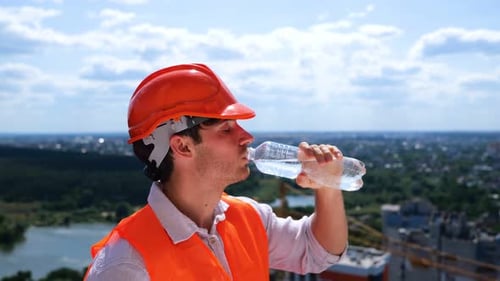 Male Builder in Helmet Standing on the Roof on the Construction Site and Drinking Water Side View