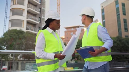 Two diverse Professionals in hard hats and vests reviewing blueprints near crane on building site