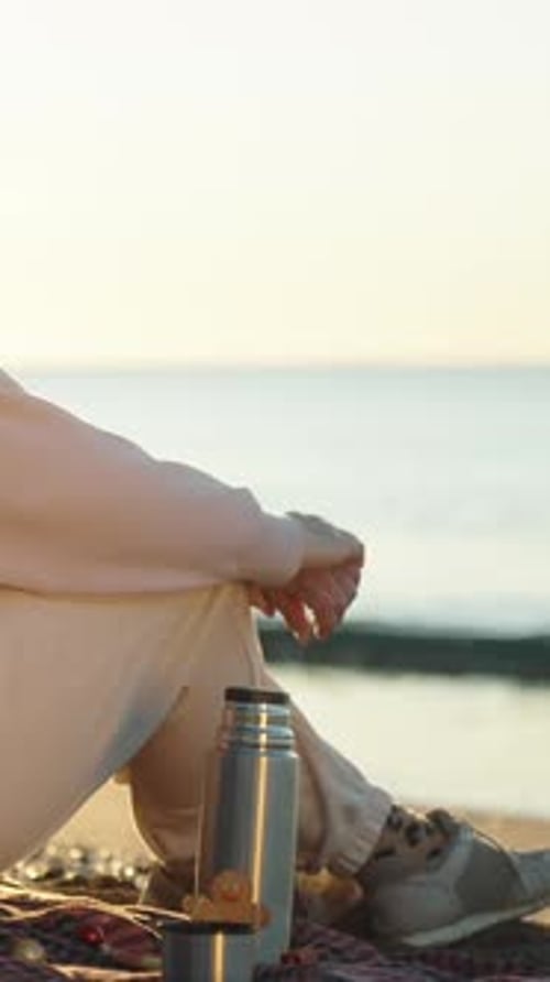 Vertical video. A young woman in a red Santa hat welcomes the sunrise by the sea. On a blanket, ther
