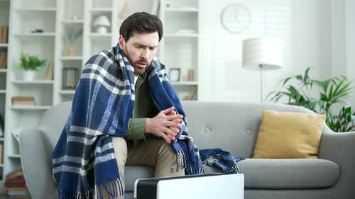 Upset man covered with a blanket frozen sitting on sofa in living room at home. Disappointed bearded