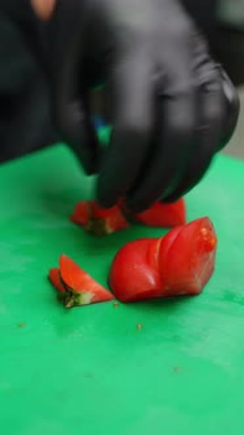 Un chef con guantes negros corta de manera experta un tomate rojo maduro en trozos precisos sobre un verde vibrante