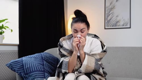 Young Woman Sits on the Couch at Home Under a Plaid Sneezing in a Napkin Feels Weak Unwell Needs