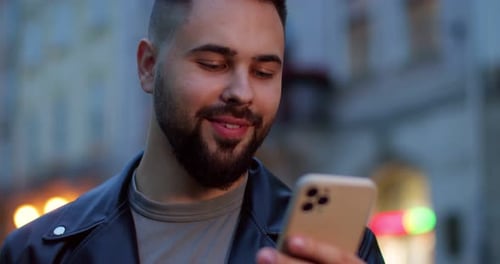 Close Up of Cheerful Young Handsome Smiling Male in Leather Jacket Texting Sms on Cellphone and