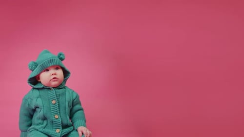 Portrait of a Cute Baby in a Warm Knitted Jumpsuit with a Hood on a Pink Background