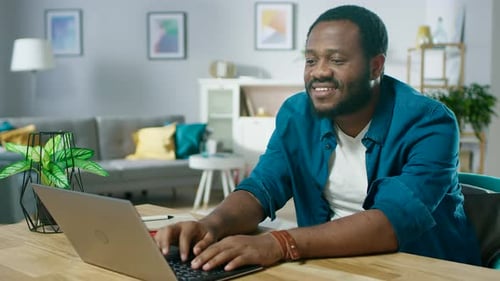 Bonito homem negro sorridente usando laptop enquanto está sentado na mesa de sua aconchegante sala de estar.