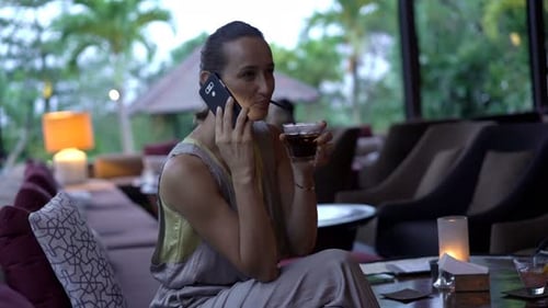 Woman Talking On Cellphone And Drinking Beverage Sitting In Garden In Cafe