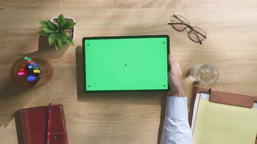 Top down view of man's hand holding a green screen tablet on a wooden desk