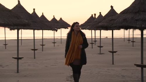 Young lady enjoying tranquility of empty beach walking through straw umbrellas on sand