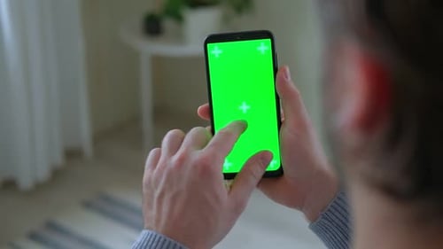 Young Man Sitting at Home Holding Smartphone Green Mockup Screen in Hand Male Person Using Chroma