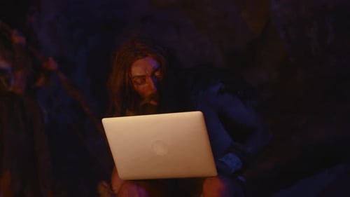 A Caveman Humorously Using a Modern Laptop in a Primitive Setting Creates an Amusing Contrast