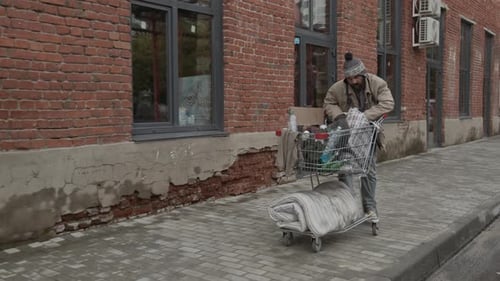 Poor Mature Man Riding Shopping Cart with His Belongings down Street