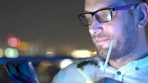 Handsome young man browsing smartphone and drinking cocktail at rooftop bar at night