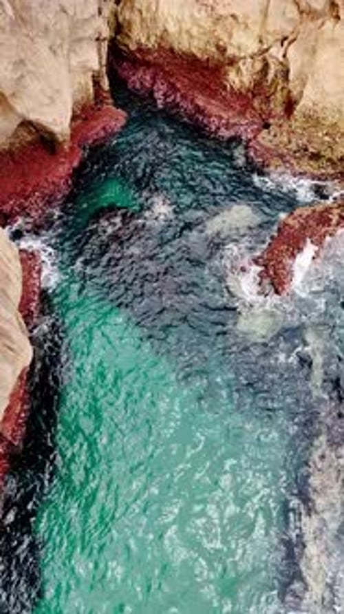 Sea water splashes among the cliffs. Foamy waves reach the red rock formations at the shore.