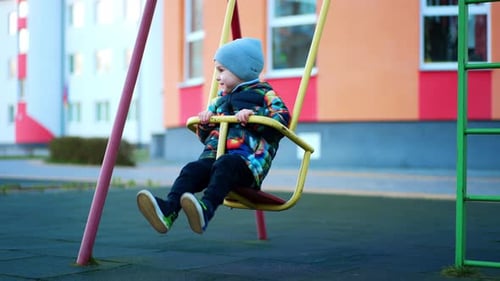Smiling baby boy in warm clothes sits in a swing outdoors. Happy child on the playground.