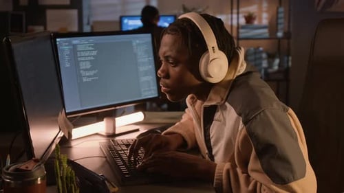 Schwarzer Softwareentwickler trägt Kopfhörer und codiert nachts am Computer